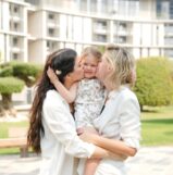 Two women kissing a smiling child in a park.